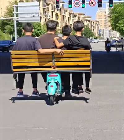 这是一张充满趣味的视觉错位照片，在城市的十字路口处，一辆浅绿色电动车后方固定着一张木质长椅，四名男生坐在长椅上。从背面视角看去，仿佛四人的身体和电动车融为一体，呈现出“悬浮”在电动车后方的错觉，看起来像是电动车违规搭载多人还加装了长椅，画面滑稽又充满猎奇感，让人第一眼产生误解。