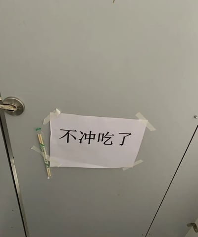这是一张实拍的公共厕所隔间场景图，浅灰色的卫生间隔间门板上，用四段透明胶带横向固定了一张白色打印纸，纸上印有四个醒目的黑色大字“不冲吃了”，在打印纸的左侧还额外用胶带固定了一双未拆封的一次性竹筷。结合厕所的使用场景，这张标语是用恶搞玩梗的方式警示如厕者使用完厕所后要冲水，否则就要承担调侃性质的“吃排泄物”的后果，搭配筷子的布置进一步强化了恶搞效果，趣味感十足。