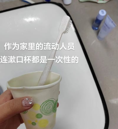 这是一张实拍的生活类图文图，画面左下角露出一只做了粉色带闪钻美甲的手，正握着一个印有黄绿螺旋圆圈图案的一次性纸杯，纸杯里插着一支白色普通牙刷，背景是带黑边的白色洗手台面，台面上还摆放着护肤品瓶罐等物品。图片上方叠加有两行白色文字：“作为家里的流动人员 连漱口杯都是一次性的”，以自嘲的口吻调侃自己在家没有固定的专属洗漱用品，处境如同临时居住的流动人员，很容易引发在外工作偶尔回家、在家没有专属个人物品群体的共鸣。