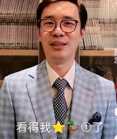 图片中是一位戴着眼镜、穿着浅蓝色格子西装的男子，内搭白色衬衫，系着带白色圆点的深色领带，西装口袋里还插着一块深色方巾。他面带微笑，背景是一排排整齐排列的书籍。图片底部有一行文字：“看得我⭐🦆①了”，其中包含星星和鸭子的emoji图案。