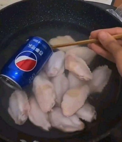 这张图片呈现了一个荒诞搞笑的烹饪场景：一口锅里装着水和多块生鸡翅，同时还有一整罐未开启的百事可乐被放进了锅中，一只手拿着筷子似乎要对锅里的食材进行操作。这是对家常菜“可乐鸡翅”的搞笑误解，正常制作可乐鸡翅是将可乐倒出作为调味酱汁使用，而非将整罐可乐与鸡翅一同下锅，这种错误操作制造出滑稽离谱的效果，让人忍俊不禁。