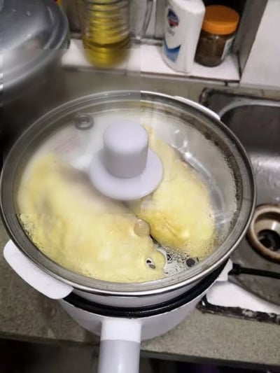 这是一张在普通家庭厨房拍摄的实拍照片，画面主体是台面上的小型电煮锅，锅内正烹煮着两块黄色的榴莲果肉，锅上盖着带白色把手的透明玻璃盖。玻璃盖的白色柱状把手恰好处于两块榴莲果肉的中间位置，和榴莲果肉、锅内的气泡组合起来，形成了酷似一张皱着眉、表情委屈的人脸的视觉错位效果，十分诙谐有趣。背景可见厨房台面上摆放的食用油、清洁用品、调料罐，旁边还有水槽区域，是典型的居家厨房环境。