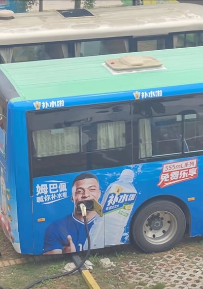 这是一张实拍的新能源公交车照片，车身印有“补水啦”电解质饮料的宣传广告，广告使用足球运动员姆巴佩作为代言人，配文“姆巴佩喊你补水啦”，旁侧还有“555mL系列 免费乐享”的促销字样。刚好公交车的充电口位置与广告上姆巴佩的嘴部位置完全重合，充电枪插入充电口的视觉效果，看起来就像是姆巴佩的嘴里插着一根管子，该巧合刚好呼应了网络上此前“姆巴佩嘴上没个把门的”的调侃，形成了极具喜感的玩梗效果。