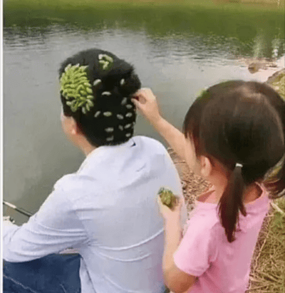 图片中一个男人和一个小女孩坐在河边钓鱼，女孩正在把一团团绿色的草或植物黏在男人的头发上，男人配合地坐着。整体画风生活气息浓厚，非常有趣，展现亲子互动欢乐时刻。画面有一点幽默和整蛊的感觉，看起来像是小朋友恶搞爸爸，充满童趣。图中没有明显的双关、谐音梗或暗示内容。