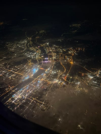 这是一张从飞机舷窗拍摄的高空城市夜景实拍图，以俯视角度呈现了夜晚的城市全貌：暖黄色的灯光清晰勾勒出城市的道路网络脉络，成片的建筑照明星罗棋布，可见穿城而过的宽阔水域，画面下方有部分云层覆盖在城市上空，左下角露出了飞机舷窗的边缘。发布者最初猜测该城市是武汉，后认为特征不符，发起无奖竞猜邀请网友判断城市身份。