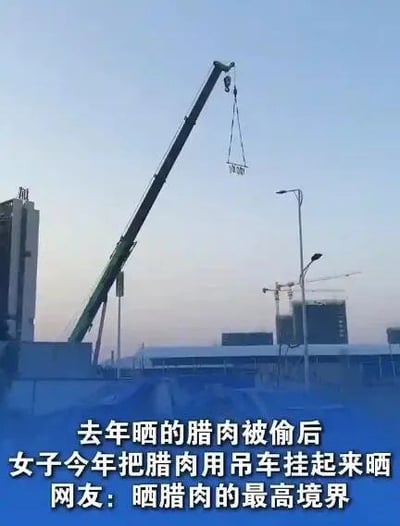 图片展示了一处建筑工地场景，一台大型吊车正吊起一个装满腊肉的篮子在空中晾晒。背景中可见高楼、路灯及其他建筑设备，天空呈现傍晚或清晨的淡蓝色。图片下方配有文字说明：去年晒的腊肉被偷后，女子今年用吊车将腊肉挂起晾晒，网友调侃这是“晒腊肉的最高境界”。整个画面因用重型机械晾晒食物的反差感显得十分滑稽，展现了民众为防盗想出的创意生活妙招。