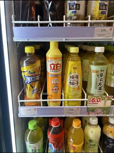 这是一张便利店冷藏饮料柜的实拍照片，柜内分层摆放着康师傅冰红茶、蜂蜜柚子茶、茉莉龙井、三得利乌龙茶、统一绿茶、阿萨姆奶茶等多款常见瓶装饮品，在第二层的饮料中间被错误放入了一瓶立白新金桔洗洁精，该洗洁精的黄橙色包装和周围同色系的饮料外观相似度较高，放在饮料陈列区极易被误认为是可饮用的饮料，形成了荒诞搞笑的反差效果。