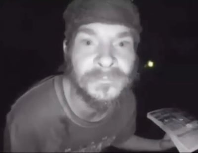 这是一张黑白夜视风格的视频截图，画面主体是一名戴着头巾、留有胡须的男子，他双眼圆睁，露出夸张的惊讶神情，身体前倾，左手拿着一个类似键盘或电子控制器的物品。背景是昏暗的环境，只有远处有一点微弱的光源，整体画面呈现出监控或夜视拍摄的效果，男子的表情极具冲击力，很适合用来表达震惊、不敢相信的情绪。