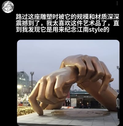 图片上方为文字描述，内容为“路过这座雕塑时被它的规模和材质深深震撼到了，我太喜欢这件艺术品了，直到我发现它是用来纪念江南style的”。下方是一张实拍照片，展示了一座大型青铜材质雕塑，雕塑主体为两只巨大的手做出手指交叉形成爱心（手指爱心）的手势，背景可见城市建筑和少量行人。整体通过文字与图像结合，形成一种因雕塑的艺术感与纪念对象（江南Style）的流行文化属性之间的反差而产生的幽默效果。