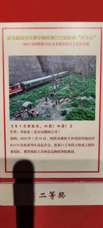 这是一幅获得2023全国铁路书法美术摄影优秀文艺作品展二等奖的摄影作品，记录了2023年7月31日的真实救援场景：因洪水滞留丰沙线沿河城站的K1178次旅客列车食品告急，张家口工务段的抢险救援队携带抢险工具与食品物资，徒步在崎岖的野外山路中向受困列车行进，列车旁的旅客也在等候救援，画面展现了铁路职工在自然灾害面前，为保障旅客安全与需求挺身而出的坚毅与暖心。