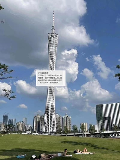 这是一张在广州塔（小蛮腰）下拍摄的实景照片，画面里蓝天白云下的广州塔矗立在城市建筑前方，下方草坪上有三名女子正躺着休息。图中添加了一段虚构的新闻文字，内容为：「广州实时新闻：2022年7月12日三名女子在广州塔暴晒，11时中暑昏迷入院，目前情况不明，后续继续更新状况，请广大市民不要盲目跟风」。这是网友制作的玩梗图，用实景配合虚构新闻来调侃夏日高温暴晒的风险，提醒大家注意防暑，没有双关、谐音梗等特殊隐含内容。