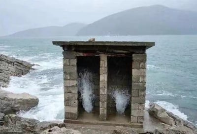 这是一张在海边礁石旁拍摄的照片，画面中的双隔间石砌小屋外观酷似户外旱厕，海浪不断涌入小屋的两个隔间，随后又从里面喷溅而出，形成了仿佛厕所被海水“使用”的滑稽视觉效果，让人脑补出在其中如厕会被海浪冲击的搞笑场景，充满了自然景观与建筑结合的反差趣味。