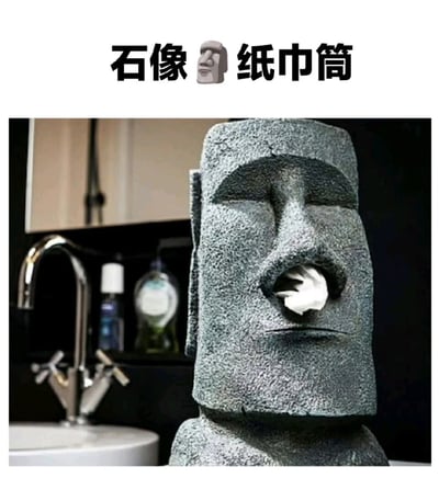 图片展示了一个设计成复活节岛摩艾石像造型的纸巾筒，放置在浴室环境中。石像面部表情严肃，嘴巴部位露出一卷纸巾，仿佛从口中吐出一般，极具趣味性。图片上方配有文字“石像🧱纸巾筒”，直接点明产品主题。背景中可见浴室的水龙头、洗手池及洗漱用品，增强了场景的真实感。