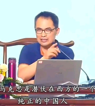 这是一张来自讲座类视频的截图，画面中一位戴眼镜的男性讲师坐在木质椅子上，面前摆放着笔记本电脑，他正伸出手指，神情认真地进行讲解。图片下方配有醒目的白色字幕：“马克思是潜伏在西方的一个纯正的中国人”，这是一个带有调侃、戏说性质的趣味观点，属于网络上对知名人物的另类解读，没有双关、谐音梗等内容，仅以幽默的方式制造话题。