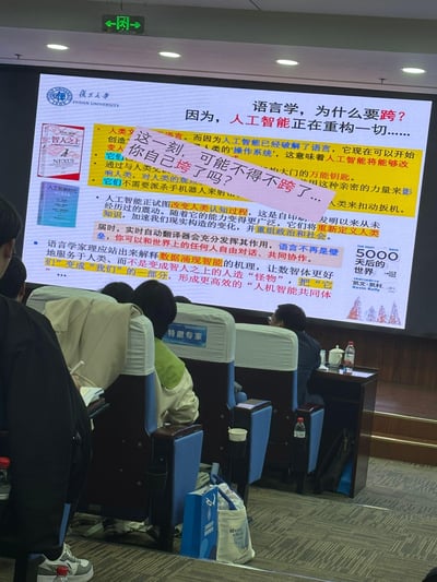 这是一张复旦大学学术讲座现场的实拍图，画面核心是讲台的大屏幕，展示着以「语言学，为什么要跨？因为，人工智能正在重构一切……」为主题的PPT。PPT内容围绕AI对语言学乃至人类社会的冲击展开：提到AI已破解语言这一人类的「操作系统」，将改变人类认知过程、重组政治社会，实时翻译将打破语言壁垒；同时呼吁语言学家研究数据涌现智能的机理，避免AI成为「智人之上的怪物」，构建人机智能共同体，还附带了《智人之上》《人工智能时代》《5000天后的世界》三本书的封面。此外，屏幕上被添加了粉色的覆盖文字：「这一刻，可能不得不跨了...你自己垮了吗？」，玩了「跨（跨界）」与「垮（处境窘迫）」的谐音梗，调侃语言学专业在AI浪潮下的处境。画面下方可见讲座现场的座椅、听众，还有一位标注「特邀专家」的参会者，整体是严肃学术场景与玩梗文字的反差感。