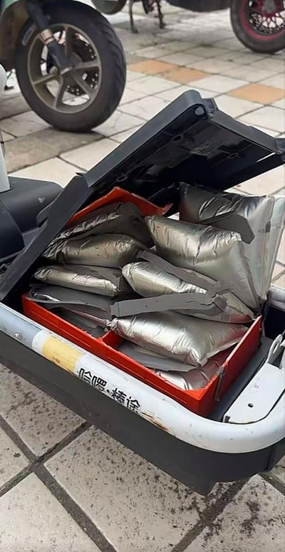 这是一张室外实景拍摄的照片，画面主体是一辆掀开了坐垫的电动车，坐垫下方的电池仓内的多块银色封装锂电池全部出现了严重的鼓包膨胀现象，变形的电芯将整个电池仓挤得满满当当，随时存在起火爆炸的安全隐患。背景可见铺设了黄灰相间地砖的路面，以及其他车辆的轮胎，电池仓外壳上有反向印刷的模糊中文字样无法辨认内容。