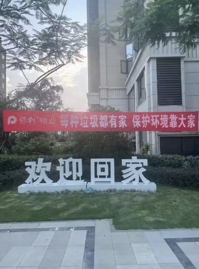 这张实拍图展示了新交付住宅小区的入口景观，前景是灰色拼花步道砖，步道后是一片草坪，草坪上摆放着四个大型白色立体汉字“欢迎回家”，立体字上方悬挂着一条保利物业的红色宣传横幅，横幅左侧印有保利物业品牌logo以及“保利物业”字样，横幅主体宣传标语为“每种垃圾都有家 保护环境靠大家”。两条标语上下摆放，内容巧合组合形成了歧义性的搞笑效果，调侃意味近似“欢迎垃圾回家”，背景可见小区高层住宅楼、树木与灌木丛，整体是真实的小区实拍场景。