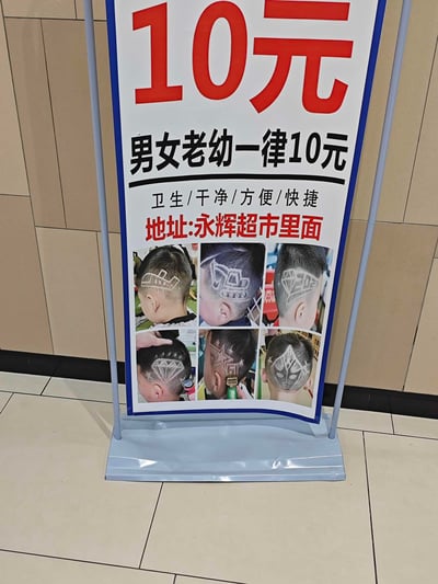 这是一款线下立式理发宣传展架，主打男女老幼理发一律10元的低价服务，宣传卖点为卫生、干净、方便、快捷，店铺地址位于永辉超市内部。展架下方展示了6款儿童创意雕刻发型的实拍图，包含汽车、钻石、面具等趣味图案，以此展现店铺的创意理发能力，吸引不同年龄段的顾客，尤其是有儿童理发需求的家长。