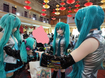 这是在国内同人漫展现场拍摄的画面，三位身着初音未来同款公式服、佩戴kigurumi头壳的爱好者聚集在一起互动，其中一人抱着初音未来的毛绒玩偶，一人手持标志性的葱形道具，周围是其他参展的人群，天花板悬挂着红黄相间的灯笼，现场氛围热闹温馨，呈现出虚拟角色“线下聚会”的趣味场景，没有双关、谐音梗或暗示内容。