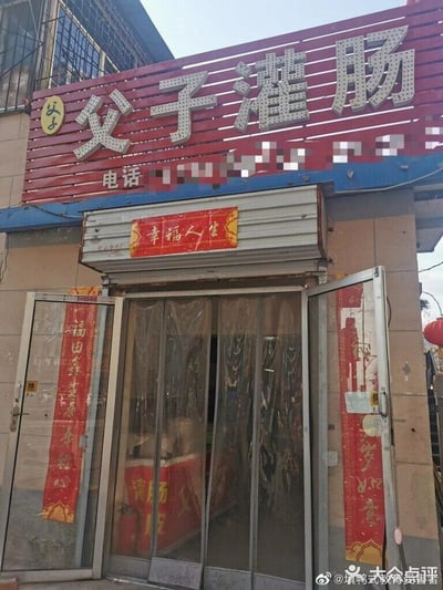 这是一张街边小吃店「父子灌肠」的门头实拍图，店铺采用红底白字的醒目招牌，清晰标注店名，招牌下方的联系电话被打码处理。店门处悬挂着红色新春横批与对联，横批为「幸福人生」，左侧对联写着「福由善生春常驻心」，右侧对联为「岁岁如意」；店门安装了塑料门帘，门内的柜台也印有「灌肠」字样的标识，整体风格接地气，充满市井烟火气，是一家主打灌肠类传统小吃的个体店铺。图中无谐音梗、双关或暗示类内容。