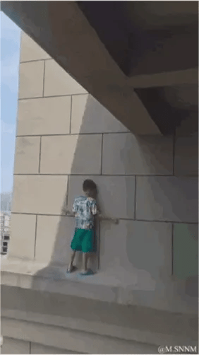 这是一则利用建筑结构制造视觉错觉的动态GIF图。画面中的小男孩看似站在高层建筑的外沿平台上，仿佛身处危险的高空险境，让观看者产生惊险的感受；但实际上是拍摄视角造成的错觉，小男孩处于安全的低位平台，这种强烈的反差带来了搞笑又意外的效果。