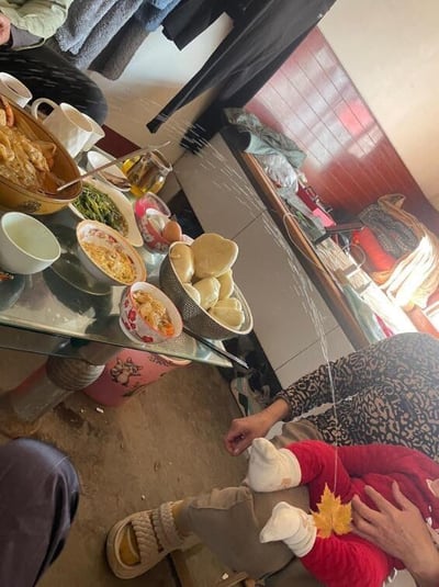 这是一张充满生活趣味的家庭实拍照片，场景是室内的餐桌旁，桌上摆放着馒头、汤面、清炒时蔬等丰盛的家常菜肴，看起来是正在进行的家庭聚餐。画面下方，一位大人抱着身穿红色衣服的小宝宝，利用巧妙的视觉错位，让一片黄色枫叶刚好位于宝宝的屁股位置，还手绘了白色线条模拟“尾巴”的形态，让枫叶看起来像是宝宝长出来的小尾巴，制造出温馨又搞笑的可爱效果，充满了家庭生活的小乐趣。