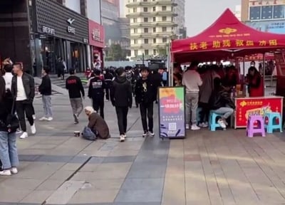 这是一张横版的城市商业步行街实拍图，展现了热闹的街头场景。画面左侧靠近边缘处，一位白发老人坐在铺着地砖的地面上，身旁放着背包，面前有一个容器，呈现出乞讨的状态。画面中间及左侧区域有多名穿着休闲服饰的年轻人，正沿着步行街行走。画面右侧是一个红色的中国福利彩票临时摊位，帐篷上方印有“扶老助残 救孤济困”的字样，摊位前聚集了不少人，旁边的立牌写着“99公益日 一块做好事”的公益宣传语。背景中可以看到“ERKE（鸿星尔克）”的品牌门店，远处还有一栋具有多层塔楼结构的特色建筑，整体场景充满了城市商圈的烟火气，同时也呈现出乞讨者与公益摊位共存的现实画面。