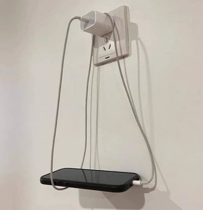 图片展示了一个白色墙壁插座，上面插着一个白色充电器，充电器连接的灰色充电线被巧妙地弯曲成弧形，下方悬挂着一部黑色智能手机，手机通过充电线的张力悬空固定，形成一种创意的充电支撑方式。整个场景简洁，背景为纯白色墙壁，突出展示了利用充电线临时支撑手机的生活小技巧。