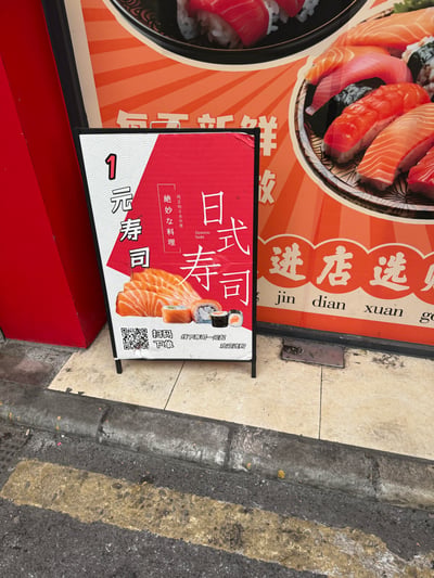 这是一张在街边寿司店门口拍摄的宣传立牌，立牌以红白配色为主，搭配三文鱼寿司、多款卷寿司的实物图，主打「1元寿司」的低价卖点，同时提供扫码下单的便捷渠道。立牌后方是店铺的大型寿司宣传海报，整体物料用于吸引路过的行人，引导大家到店选购寿司，没有双关、谐音梗或隐含暗示内容。