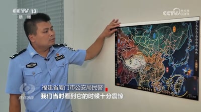 画面中一名身穿蓝色警服的福建省厦门市公安局民警，正站在一面墙前，用右手指着墙上的一张复杂地图。民警表情严肃，背景中能看到百叶窗。屏幕左上角有“CCTV 13 新闻”标识，右上角有“CCTV.com”水印，底部左侧显示“焦点访谈”字样，底部中间有字幕“我们当时看到它的时候十分震惊”。