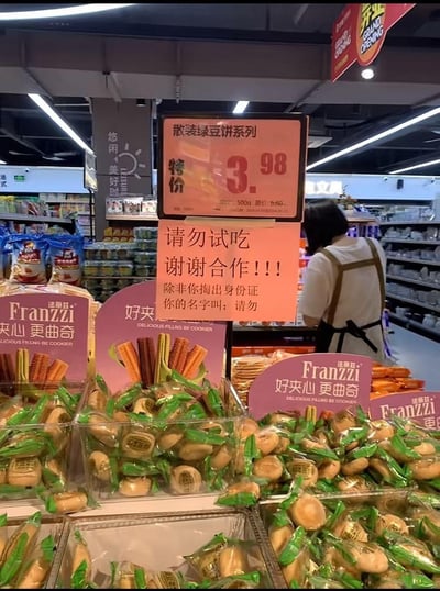 超市零食区场景，货架上摆放着法丽兹（Franzzi）夹心曲奇等商品，最显眼处是粉色的散装绿豆饼系列特价牌，标注'3.98元/500g（原价6.50元）'。价格牌下方有一张手写补充牌，原本严肃提示'请勿试吃 谢谢合作！！！'，却突然反转：'除非你掏出身份证 你的名字叫：请勿'，用谐音梗和角色扮演的幽默方式化解了警示牌的生硬感，让人会心一笑。背景可见超市天花板的灯带、'悠闲美好时'的区域标识，以及一位穿着白色上衣和棕色围裙的超市员工正在整理货架。