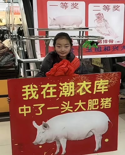 整张图为线下服饰店抽奖活动的现场实拍，画面中央是一名未成年小女孩，她穿着黑红拼接的冬装外套，表情略带局促哭笑不得，双手举着一块红色的中奖宣传牌，牌上印着白色大肥猪图案和黄色文字「我在潮衣库 中了一头大肥猪」，她的身前还放着一团红色的颁奖绸布。背景处的活动公示展板显示本次活动一等奖是整头活猪，二等奖为1扇猪排骨，背景还能看到店铺内的衣物货架等陈设，整体氛围荒诞又有喜感，小女孩的状态呈现出对中了活猪这个奖项的无措感。