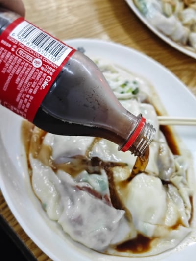 这张图里，一只手正在把一瓶可口可乐倒在一盘水饺上，看起来像是要用可乐当蘸料。画面颇有反差感，容易让人会心一笑。正常情况下水饺会蘸酱油或醋，但这里直接用可乐，不仅暗示可能是“搞怪吃法”，也带有一点双关：可乐颜色和酱油类似，容易误导。此梗图常见于展示奇葩用餐方式、或戏谑食品搭配，带有强烈的搞笑和无语气息。可能引发“为什么要这么吃”或“疯了吧”等评论。

暗示：可乐颜色像极了酱油，属于视觉错觉、反转式幽默。