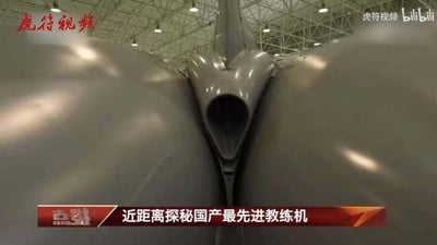 这是一张来自军事科普节目的视频截图，画面核心是国产最先进教练机的机身尾部特殊结构特写，该结构呈现出对称的流线型设计，中间有类圆形的开口，推测为进气或排气相关部件。背景是室内机库的钢结构顶棚与嵌入式照明灯具，整体环境为专业的飞机存放维护空间。画面底部带有红色字幕条，标注文字“近距离探秘国产最先进教练机”，明确了视频的主题内容。图中不存在双关、谐音梗或暗示类内容。