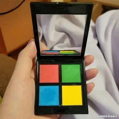 这是一张实拍静态图，画面中一只手正举着一个打开的黑色外壳四色眼影盘，眼影盘的四个色块分别为左上红色、右上绿色、左下蓝色、右下黄色，配色与微软Windows系统的经典四色徽标完全相同，眼影盘上盖的内侧自带镜子，背景能看到模糊的浅色衣物和棕色家具，整体是网友分享的趣味彩妆产品。