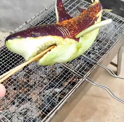 图片展示了一个创意十足的烧烤场景：一只紫色茄子被巧妙雕刻成鲨鱼的形状，正在烤架上烧烤。茄子的头部被精心雕琢出鲨鱼张开的嘴巴，露出锯齿状的“牙齿”，身体部分保留了茄子的紫色表皮，上面撒满了红色的辣椒粉作为装饰和调味。一只手用筷子从鲨鱼嘴部穿过，将这个创意作品固定在烤架上。烤架下方可以看到燃烧的炭火，整个画面展现了民间美食创意的趣味性。