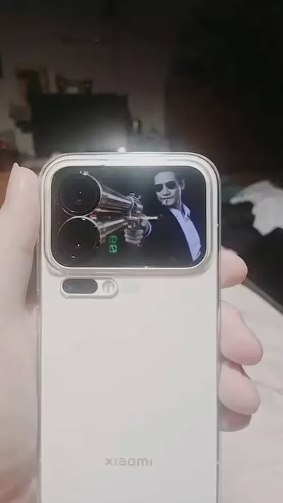 一只手握着白色小米手机，手机背面的后置双摄镜头与屏幕中显示的西装男子手持双枪画面完美对齐，镜头恰好位于枪管口位置，形成视觉错位效果，看起来仿佛手机摄像头变成了双枪的枪口，创意十足又带点酷炫搞笑。