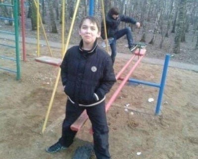 一名男孩双手插兜站在 playground 中，身着黑色夹克与黑色长裤，表情平静。其身后有另一名男孩正坐在红色跷跷板上。背景可见黄色秋千、绿色攀爬架等游乐设施，周围是树木与泥土地面，整体呈现出户外 playground 的日常场景，氛围 casual 且 candid，像是童年生活中的抓拍瞬间。