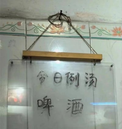图片中是一块悬挂在瓷砖墙上的玻璃写字板，瓷砖边缘带有粉色花卉与绿色藤蔓图案装饰。写字板通过木质横杆和铁丝悬挂于天花板，板上手写黑色字迹内容为“今日例汤”与“啤酒”，将通常作为饮品的啤酒列为“例汤”，形成明显的品类错位，营造出幽默搞笑的效果。场景疑似小型餐馆或食堂的内部墙面，整体环境带有生活化的朴素感。