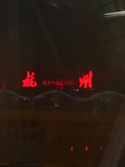夜晚拍摄的杭州地标场景，画面中央为红色发光的“杭州”汉字及下方的“HANGZHOU”英文标识，标识下方有蓝色波浪形灯带装饰。照片可能透过玻璃拍摄，存在轻微反光和模糊效果，底部隐约可见“AIR”字样，整体呈现城市夜景的灯光氛围。