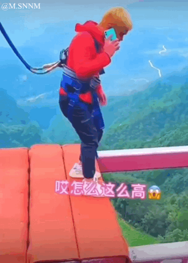 图中是一个穿着红色上衣、蓝色蹦极装备的人站在蹦极高台边缘，一只手拿着手机贴在耳边，身体姿态紧绷，看起来非常紧张害怕，下方配文“哎怎么这么高”，背景是郁郁葱葱的山间景色，整个场景是蹦极前的瞬间，人物的害怕和蹦极的刺激形成反差，十分搞笑。
