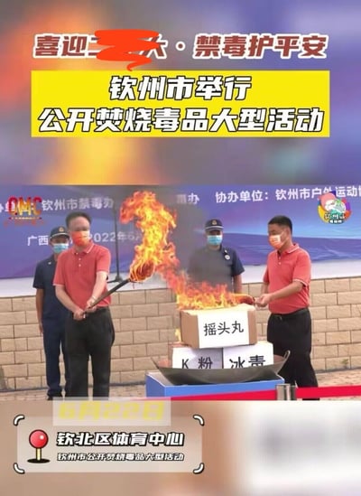 图片展示了钦州市公开焚烧毒品大型活动的现场场景。背景横幅写有“喜迎二十大·禁毒护平安”字样，黄色主标题区域突出“钦州市举行公开焚烧毒品大型活动”。前景中两名身着红色衬衫的人员正手持火炬点燃堆放的毒品箱，箱子分别标注“摇头丸”“K粉”“冰毒”，火焰正在燃烧。周围有身着制服的工作人员站立，底部标注活动时间为6月22日，地点在钦北区体育中心。