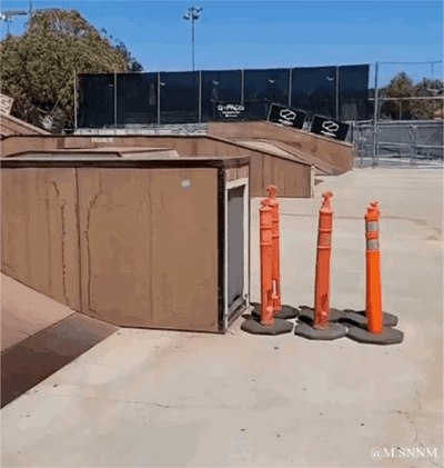 这是一段实拍的动态图（GIF），呈现了滑板场地里的趣味视觉错觉：画面中的橙色路障柱在特定视角下，刚好和旁边的棕色箱子门位置重合，看起来仿佛是从箱子门里延伸出来的，制造出荒诞又有趣的视觉效果，让人产生柱子“生长在箱子内部”的错觉。