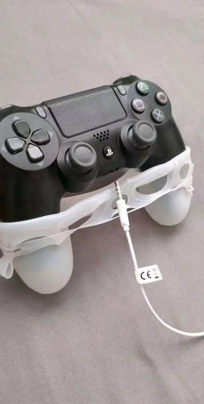 一张展示PlayStation 4手柄的照片，手柄底部被加装了一个白色的塑料/泡沫配件，看起来像是某种自制的改装。配件上连接着一根白色的线缆，线缆上有一个带有CE认证标志的标签。整个装置放在灰色的平面上，手柄保持着正常的姿态，但底部的改装配件显得有些突兀和不寻常。