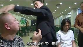 图片场景发生在一个有吊顶灯的餐厅或食堂内，一名身穿黑色上衣的男子正扬起右手，做出要打人的动作。他面前坐着一位光头男子，仰头注视着他，表情略显紧张；旁边还有一位穿白色上衣的男子低头看向桌面。图片底部有一行字幕文字：“给你机会你不中用啊”。