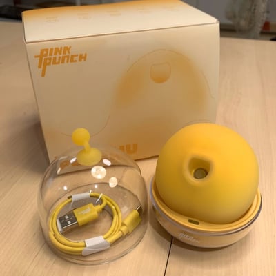 图片展示了PINK PUNCH品牌的黄色球形产品及其包装和配件。背景是一个浅黄色渐变的产品包装盒，上面印有品牌名称"PINK PUNCH"和一个抽象的圆形图案。前景中有两个主要物品：左侧是一个透明的圆顶形保护罩，里面整齐地放置着一根黄色的USB数据线；右侧是一个黄色的球形主体产品，顶部有一个圆形开口，放置在一个配套的黄色底座上。整个场景在浅色木质桌面上拍摄，光线明亮，产品细节清晰可见。