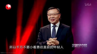 这是政论节目《这就是中国》的视频截图，主讲人张维为身着深色西装、佩戴眼镜，双手交握于身前，面带温和的笑容正在进行演讲，屏幕下方配有字幕「所以千万不要小看勇往直前的年轻人」，传递出对年轻群体的肯定与鼓励。