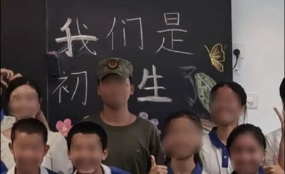 这是一张原始拍摄的静态单图合影，画面中有一位佩戴军训帽、身着深色上衣的成年人，周围环绕着多名穿着蓝白相间校服的学生，所有人物的脸部都做了马赛克处理。背景是一块黑板，上面用白色粉笔书写着“我们是初中生”，但中间的“中”字被画面中央的成年人身体完全挡住，从视觉效果上看文字变成了“我们是初生”，形成了趣味的视觉错位效果。黑板上还绘制有蝴蝶、花朵等简单的粉笔画作为装饰，部分学生做出比耶、竖大拇指的姿势，整体氛围轻松活泼，应该是新生军训期间的校园记录。
