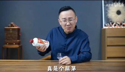 一名身穿蓝色衬衫的中年男子坐在木质餐桌前，右手持一瓶白色标签、红色瓶盖的白酒，左手握着透明玻璃杯，正处于倒酒的动作中。背景为深色墙面，左侧有雕花木箱，右侧有带抽屉的木质柜子及装饰画。图片底部有白色字幕'真是个屌茅'，右上角可见'一凌说酒 bilibili'水印，整体场景疑似酒类品鉴或评测视频截图。