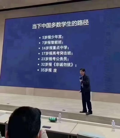 这是一张线下演讲现场的实拍图，一位男性演讲者手持麦克风站在大屏幕前进行分享。屏幕上以“当下中国多数学生的路径”为标题，列出了一系列以“报”为核心的人生节点：5岁报少年宫；7岁报智能班；14岁报重点中学；17岁报高考突击班；23岁报考公务员；32岁报《非诚勿扰》；35岁报 废。内容用“报”字串联起从童年教育到婚恋、职场的人生阶段，最后用“报废”的谐音双关，讽刺了部分人被规训、按部就班的内卷人生，直指35岁在社会竞争中陷入困境的无奈现实，引发对当代年轻人成长与生存现状的共鸣。